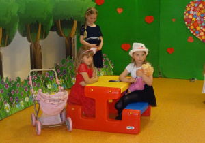 Dwie dziewczynki siedzą na ławeczce, jedna z nich trzyma lalkę. obok ławki stoi wózek. Z boku stoi dziewczynka z mikrofonem.